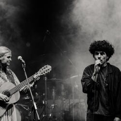 Das schwarz-weiß Bild zeigt links eine Frau mit Gitarre. Rechts einen Mann mit Mikrofon, welcher gerade singt. Die Atmospähre ist durch Nebel sehr mystisch.