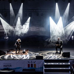 Zwei Musiker, welche mit etwas Abstand auf Stühlen sitzen. Es ist dunkel und sie werden jeweil von Scheinwerfern beleuchtet, die dann auch noch ein Muster auf den Boden werfen.