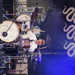 Schlagzeuger in Action. Er hält beide Drumsticks nach oben und sein Mund ist weit aufgerissen, so als würde er mitsingen.