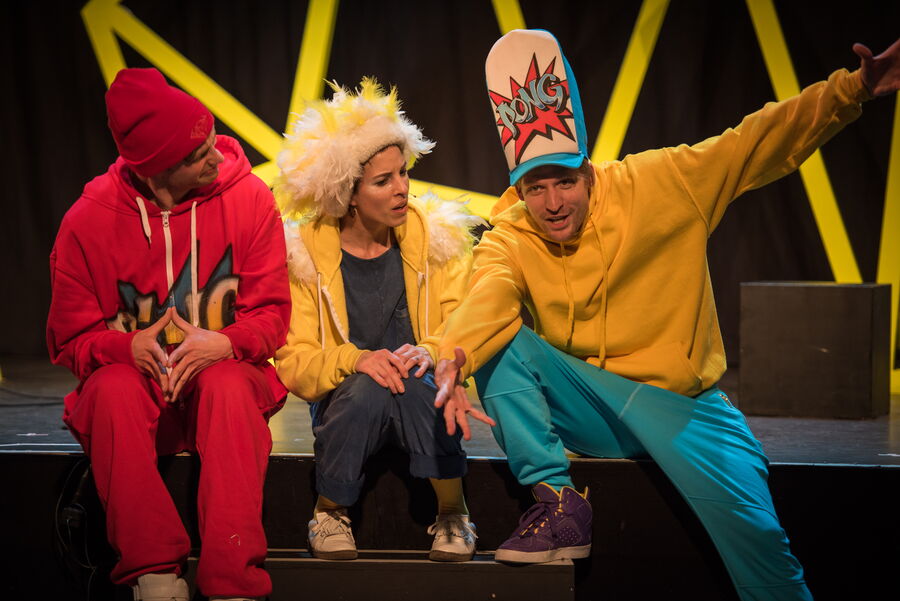 Drei Personen in bunten Klamotten sitzen auf einer Bühne. Links sitzt eine Person in einem roten Trainingsanzug und einer roten Mütze, in der Mitte eine Person in gelber Jacke mit weißem, federartigen Kopfschmuck und rechts eine Person in gelbem Hoodie, e