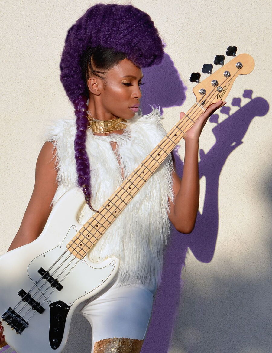 Das Bild zeigt eine Nik West mit einer auffälligen, lila gefärbten Frisur. Sie trägt ein weißes Outfit mit Fellweste, das in Kombination mit ihrem goldenen Schmuck edel wirkt. In den Händen hält sie eine weiße Bassgitarre, auf die ihr Blick gerichtet ist.