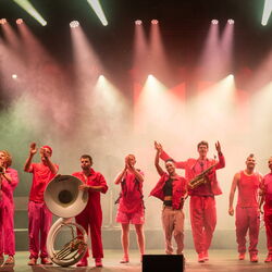 Das Bild zeigt die gesamte Band, 10 Personen. Alle tragen rote Kleidung und sind in einer Reihe aufgestellt. Sie winken und klatschen.