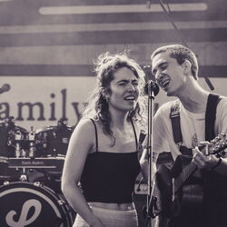 Das Bild zeigt zwei Musiker, die sich ein Mikrofon teilen. Links eine Frau und rechts ein Mann mit Gitarre.