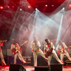 Das Bild zeigt eine Band von 6 personen alle spielen ein Instrument und sie stehen auf einer Bühne. 4 Männer sind in der gleichen Pose, un welcher sie auf dem rechten Bein stehen und das Linke in die Luft heben.