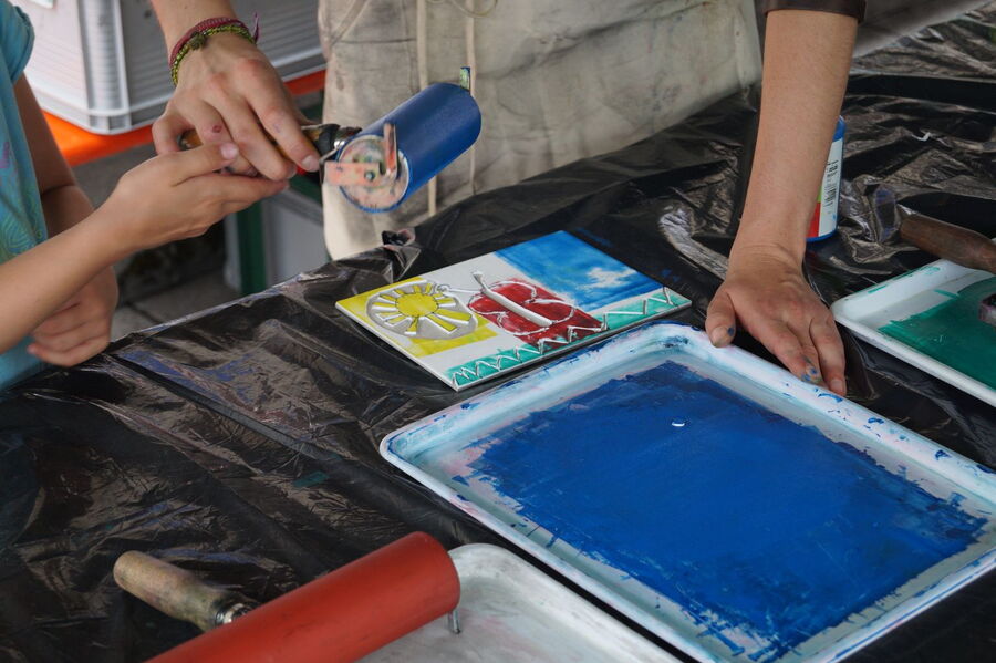 Das Bild zeigt eine Platte, auf welcher sich blaue Farbe befindet. Diese Platte liegt auf einem Tisch, davor eine Schablone mit einer Sonne und einem Schmetterling. Am Tisc stehen zwei Personen, wovon eine der anderen einen Frabroller reicht.