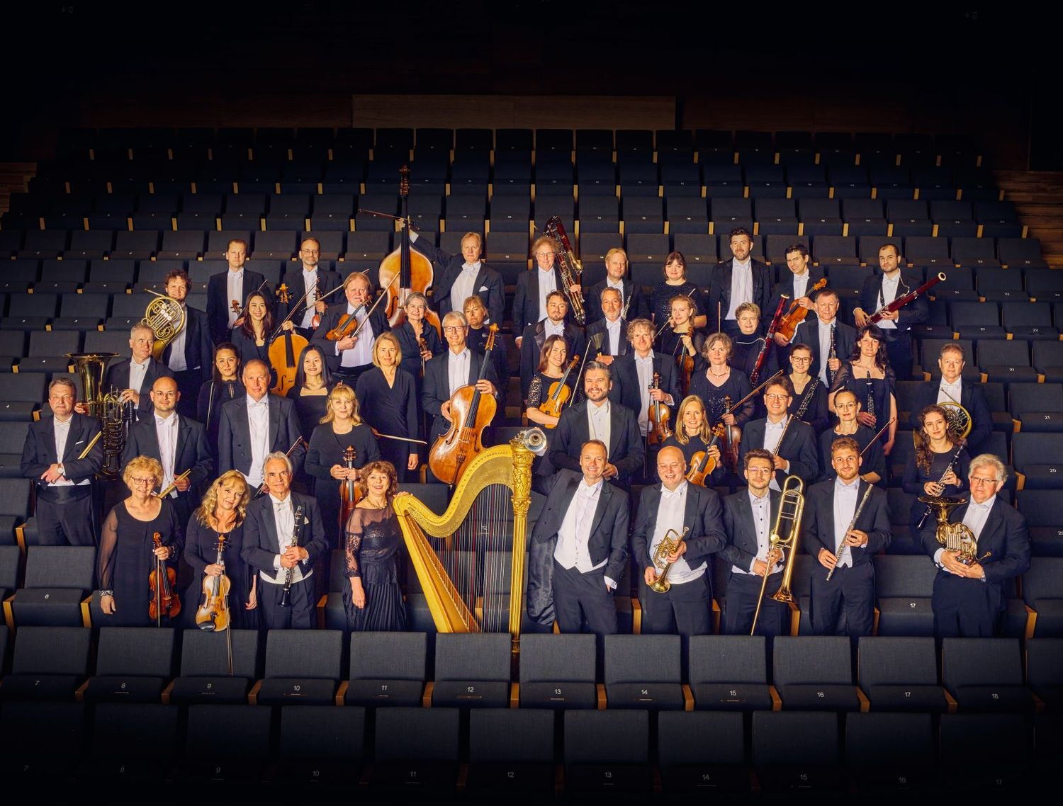 Das Bild zeigt ein Gruppenfoto des philharmonischen Orchesters Würzburg. Alle Mitglieder halten ihr Instrument in der Hand. Vorne in der Mitte sticht eine goldene Harfe hervor. Der Fokus wird durch warmes licht auf die Mitglieder gelenkt, außenrum dunkel.