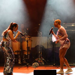 Das Bild zeigt zwei Sängerinnen, die sich anschauen und zueinander singen. Im Hintergrund sind weitere Musiker.