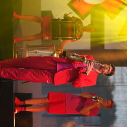 Das Bild zeigt drei Männer in roter Kleidung mit Instrumenten. Einer mit Trommel, einer mit Saxophon und der dritte mit Trompete.