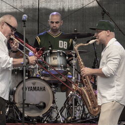 Drei Musiker, hinten der Schlagzeuger, links davor ein Posaunenspieler und rechts, ein Mann der Saxophon spielt.
