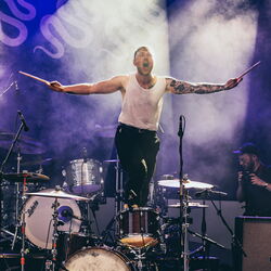 Der Schlagzeuger steht auf seinem Stuhl, er hat beide Arme mit seinen Drumsticks zur Seite ausgestreckt.
