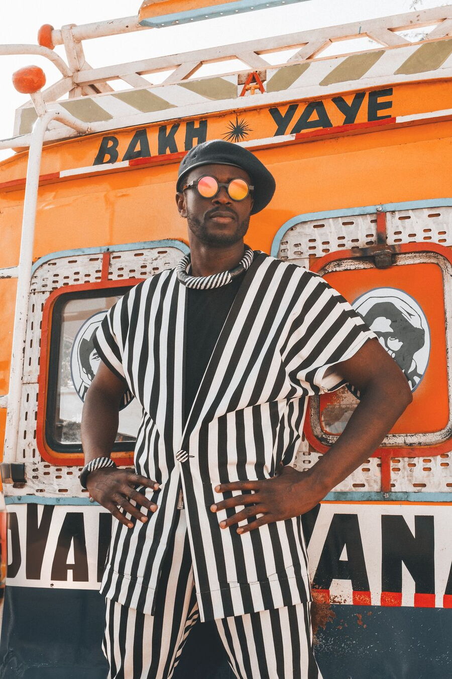 Das Bild zeigt Alune Wade vor einem orangefarbenen Fahrzeug mit auffälligem Design. Er trägt ein gestreiftes, schwarz-weißes Outfit und eine schwarze Baskenmütze. Seine Sonnenbrille reflektiert bunte Farben, was dem Bild einen kreativen Touch verleiht.