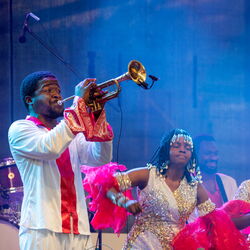 Im Vordergrund des Bildes ist ein Mann, welcher Trompete spielt. Im Hintergrund ist ein Schlagzeuger und Tänzerinnen. Alle tragen schicke weiße Kleidunf mit bunten Farbakzenten.