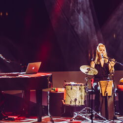 Das Bild zeigt links einen Mann welcher an einem Flügel sitzt. Rechts vom Flügel stehen viele Intrumente, vor allem Trommeln. Mitten drin sitzt eine Frau welche grade in ein Mikrofon singt.