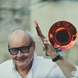 Nils Landgren hebt seine Trompete neben seinen Kopf, sein Blick ist auf den Boden gerichtet.